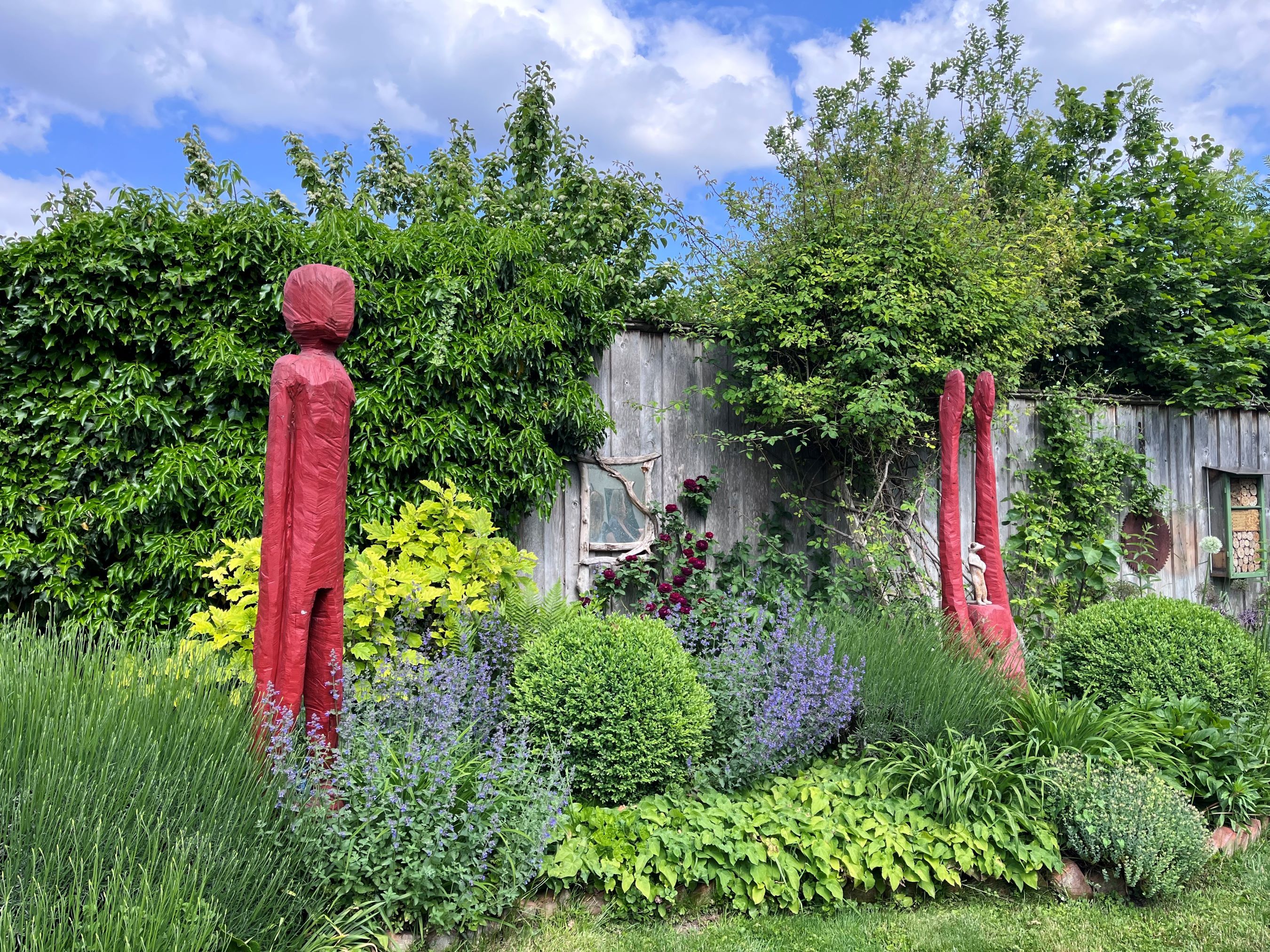 Garten mit roten Skulpturen, umgeben von grünen Pflanzen und einer Holzwand im Hintergrund.