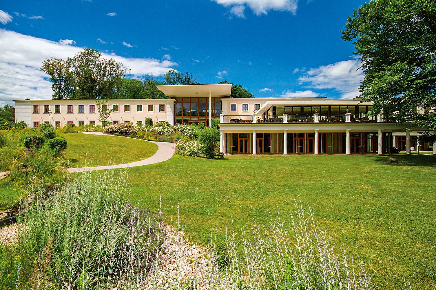 Ein modernes Gebäude mit großen Fenstern und einer Terrasse, umgeben von einem gepflegten Garten und Bäumen unter blauem Himmel.