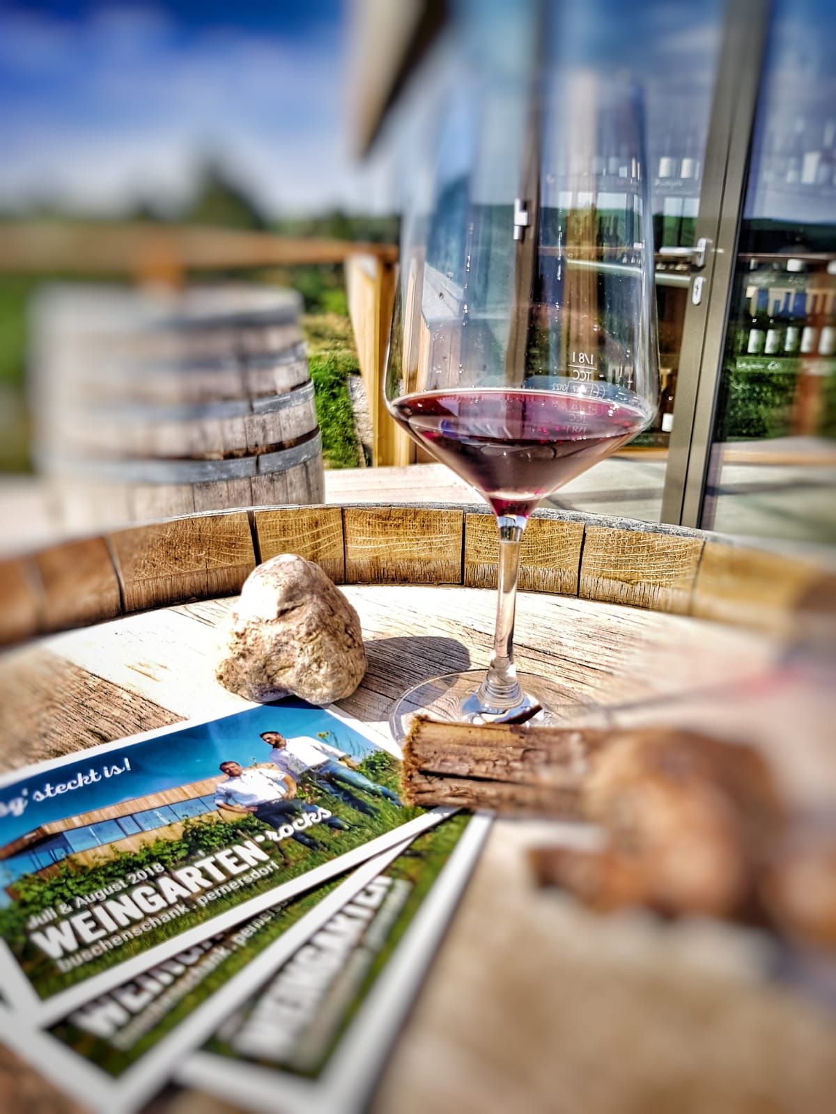 Ein Weinglas mit Rotwein steht auf einem Holztisch neben Broschüren und einem Stein.