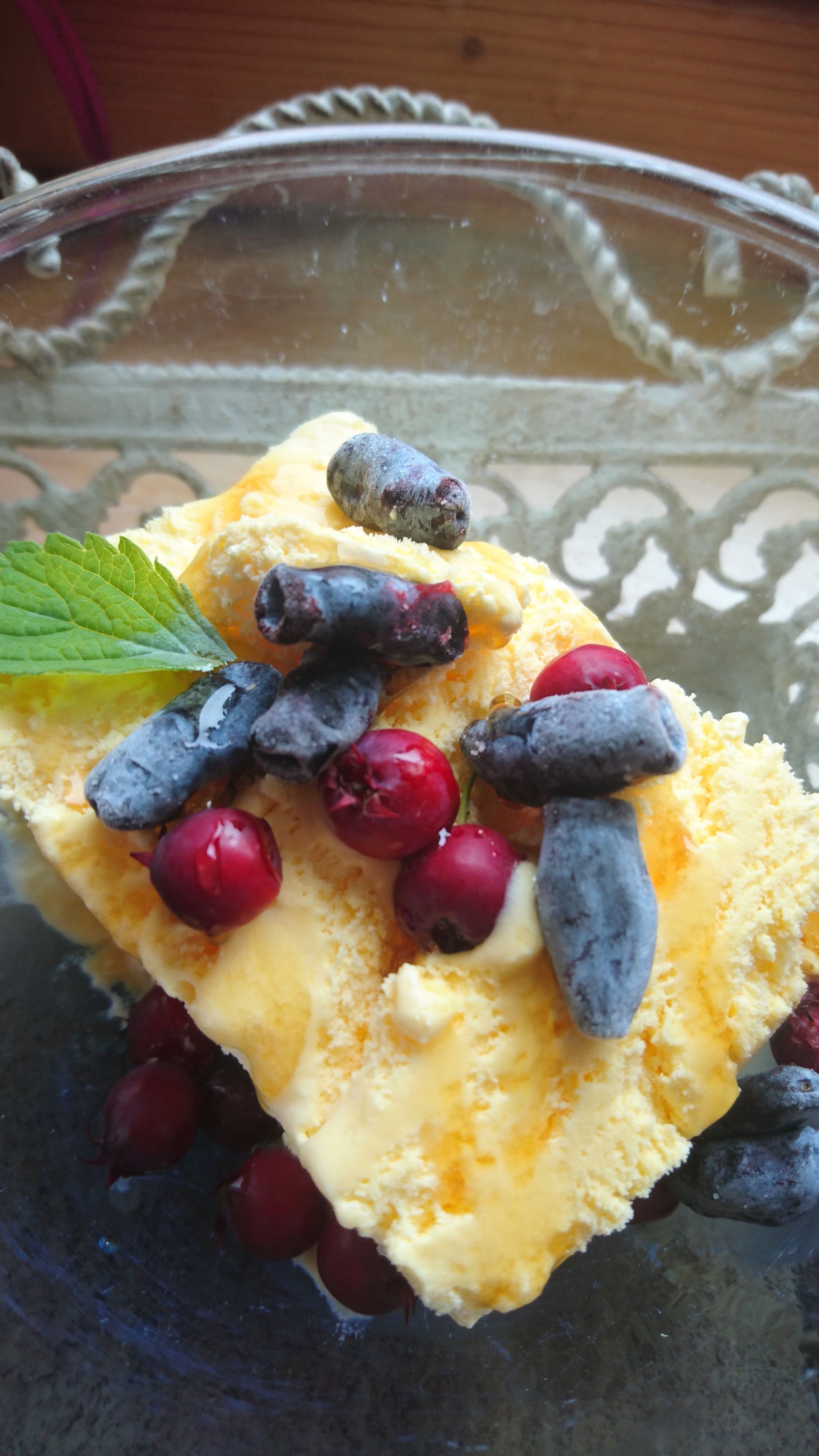 Ein Dessert mit gelbem Eis, roten und blauen Beeren, garniert mit einem grünen Blatt, in einer Glasschale.