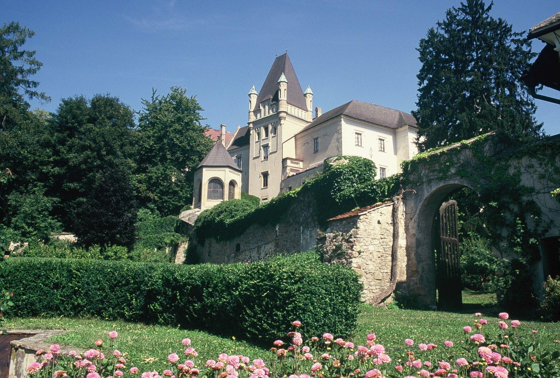 Ein Schloss mit Türmen und einer Steinmauer, umgeben von grünen Bäumen und blühenden Blumen im Vordergrund.
