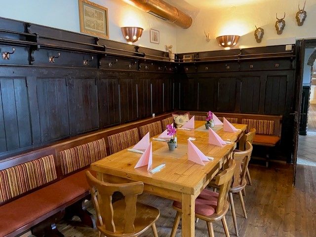 Gemütliche Gaststube mit Holztisch, Stühlen und Bank. Tisch ist mit rosa Servietten und Blumen dekoriert. Dunkle Holzvertäfelung an den Wänden.