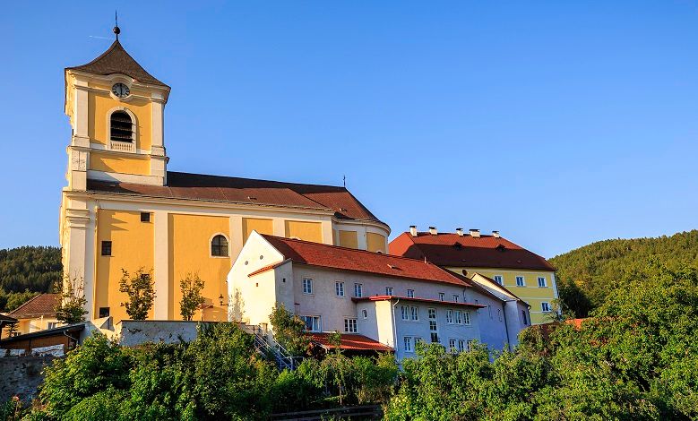 Pfarrkirche mit angrenzendem Kloster in sonniger Landschaft.