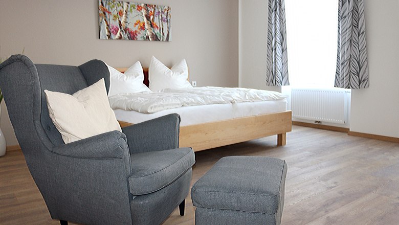 Grauer, gem&uuml;tlicher Stuhl im Vordergrund. Im Hintergrund ein Doppelbett mit wei&szlig;er Bettw&auml;sche.