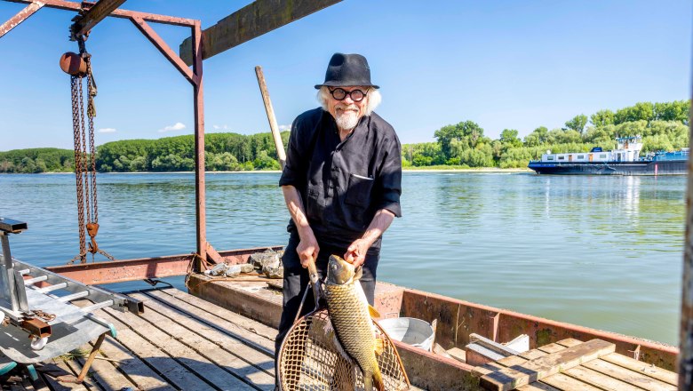 Ein Mann mit Hut hält einen großen Fisch in einem Netz auf einem Holzsteg am Fluss (Donau).