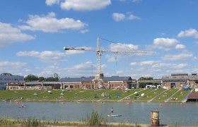 Menschen entspannen auf einer Liegewiese an einem Fluss mit einem Kran im Hintergrund.