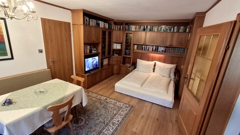 Wohnzimmer mit ausziehbarer Couch, B&uuml;cherregal, Esstisch und Fernseher.
