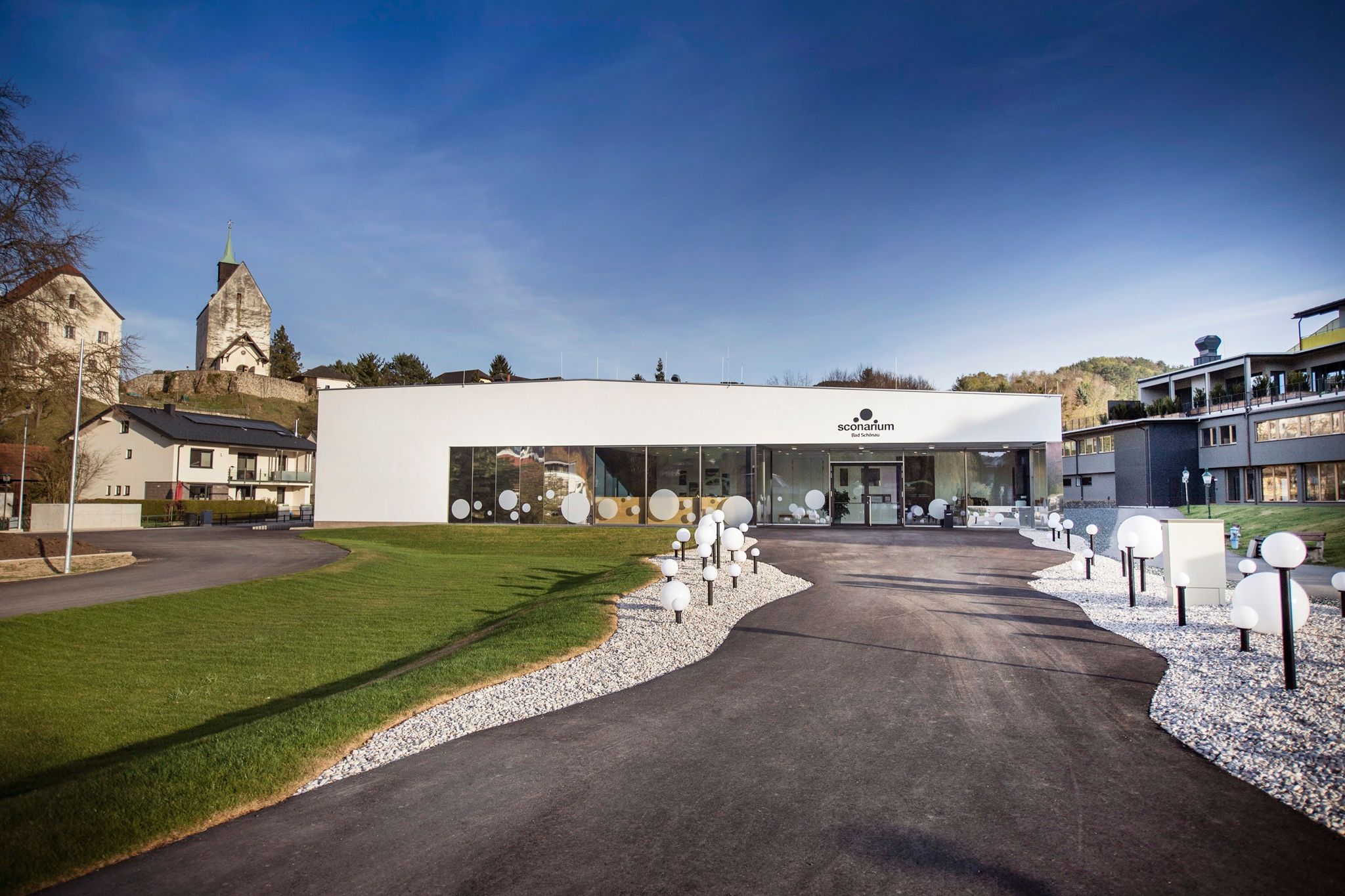 Modernes Gebäude des Sconariums mit Glasfront und dekorativen Kugelleuchten, umgeben von Grünflächen und der Wehrkirche im Hintergrund.