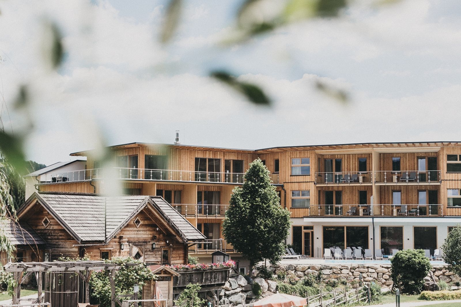 Ein modernes Holzgebäude mit Balkonen und ein traditionelles Holzhaus im Vordergrund, umgeben von Bäumen und Gartenanlagen.