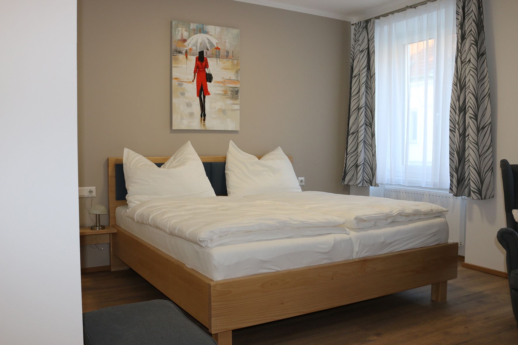 Doppelbett aus Holz mit weißer Bettwäsche. Dahinter ein Fenster mit Gardine und ein Gemälde.