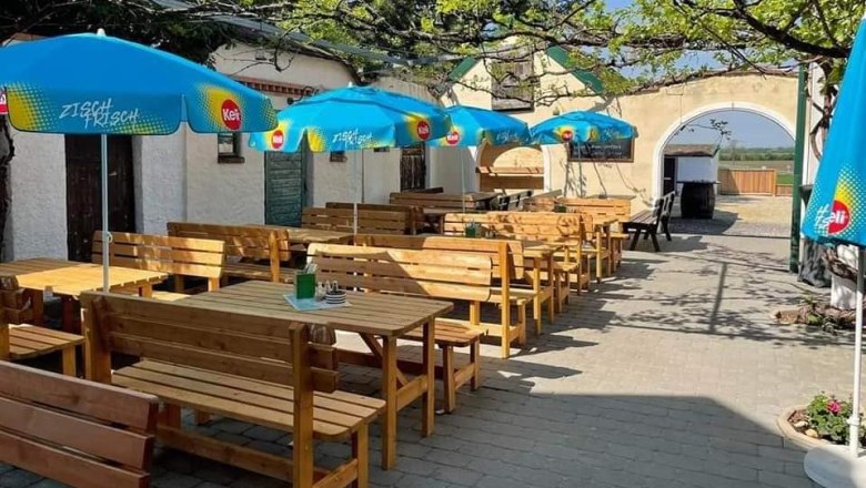 Ein sonniger Gastgarten mit Holzbänken und blauen Sonnenschirmen.