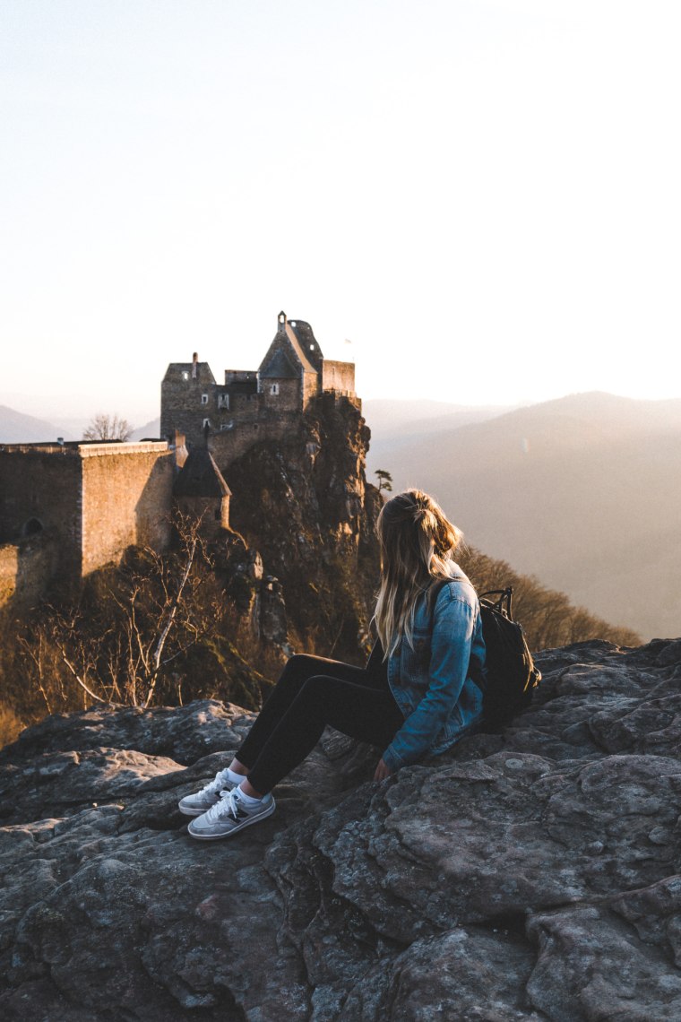 Frau sitzt auf einer alten Burgmauer und blickt auf eine alte Ruine.