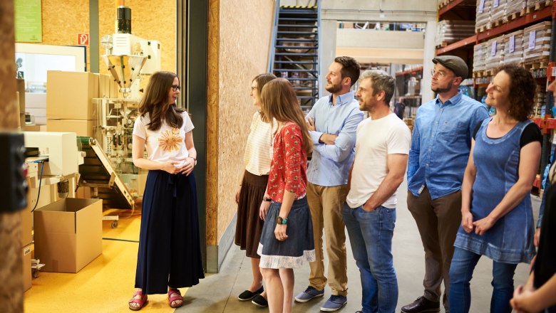 Eine Gruppe von Menschen steht in einem Lagerhaus und hört einer Frau zu, die eine Führung gibt.