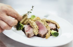 Nahaufnahme eines rosa gebratenen Fleischgerichts mit Kräuterkruste, Brokkoli und einer Hand, die Thymian hinzufügt.