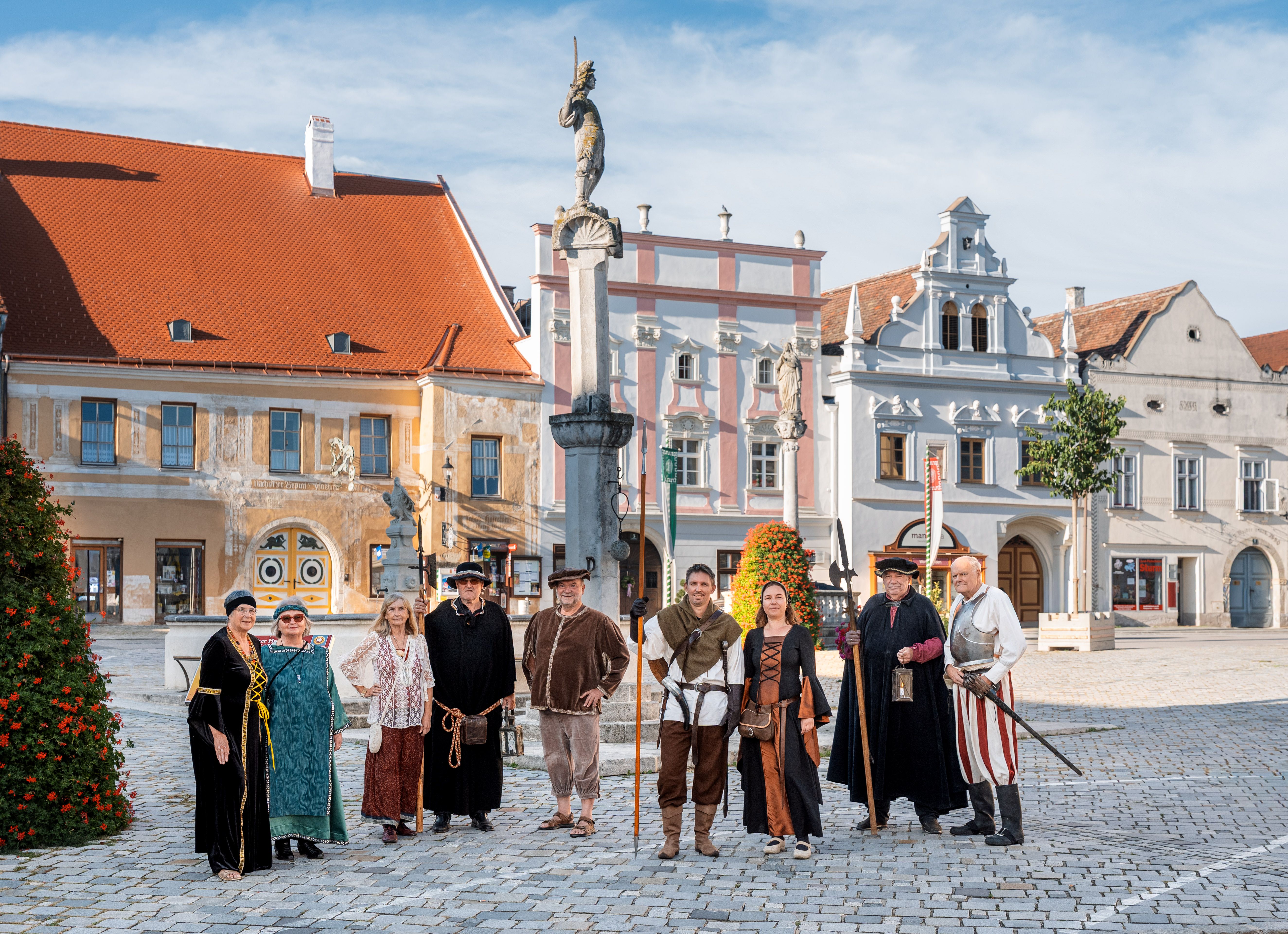 Gruppe von Menschen in historischer Kleidung auf dem Hauptplatz von Eggenburg vor barocken Häusern und einem Pranger mit Statue.