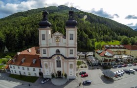 Luftaufnahme einer Kirche mit zwei Türmen, umgeben von Gebäuden und Wald.
