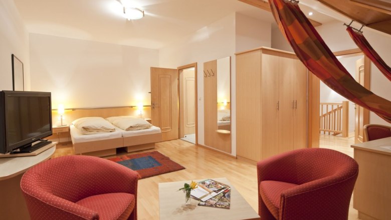 Gemütliches Gästezimmer mit Doppelbett, TV, roten Sesseln und Hängematte.