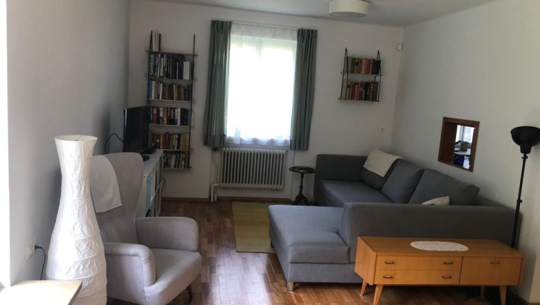 Ein Wohnzimmer mit grauem Sofa, Sessel, B&uuml;cherregalen und Holzboden.