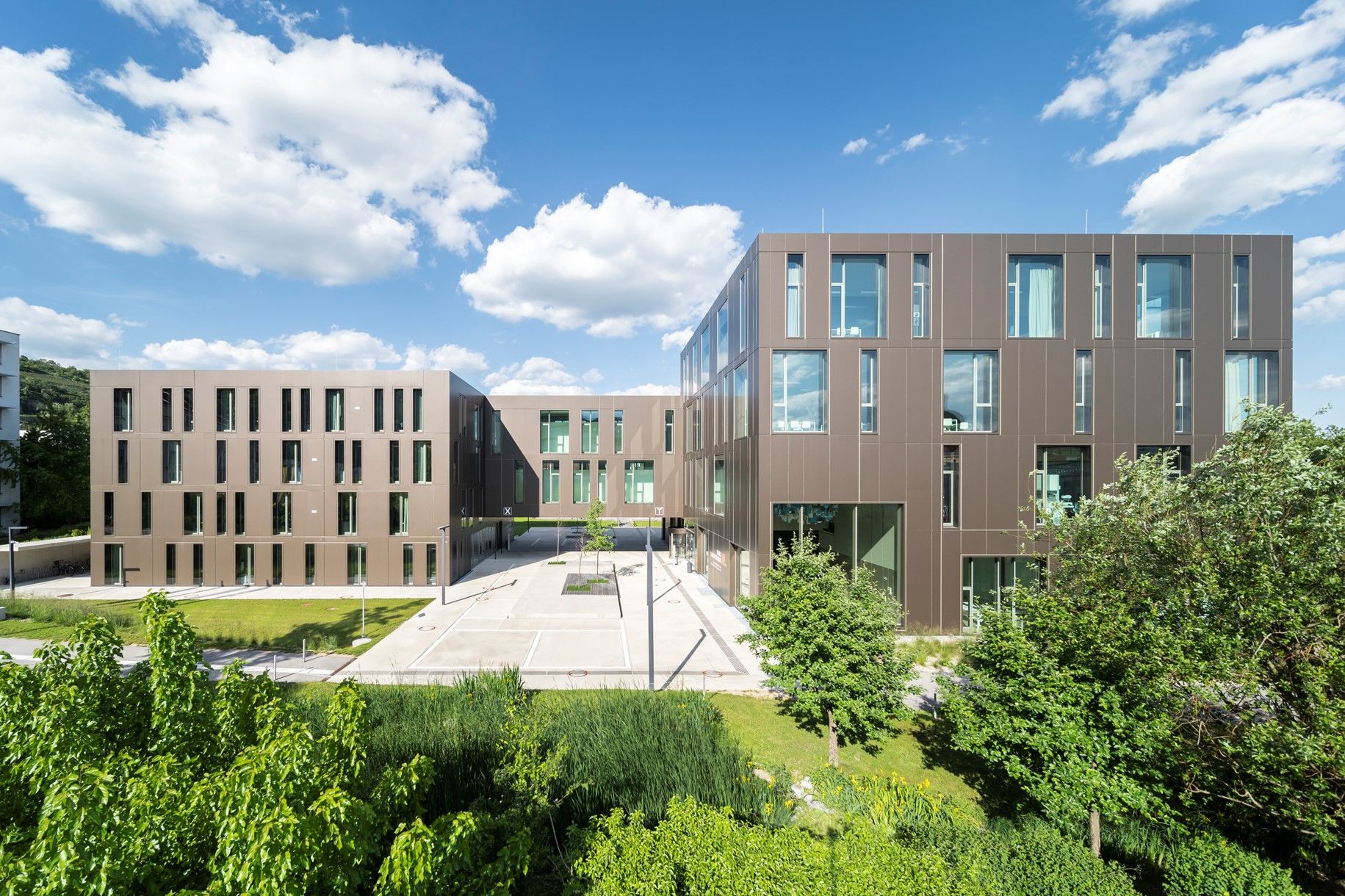 Moderne Universitätsgebäude mit brauner Fassade und großen Fenstern, umgeben von grüner Vegetation unter blauem Himmel.