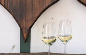 Zwei Weingl&auml;ser mit Wei&szlig;wein auf einem Holztisch vor einer Holzwand mit Herzdekor.