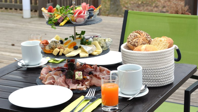 Fr&uuml;hst&uuml;ckstisch im Freien mit Aufschnitt, K&auml;se, Gem&uuml;se. Daneben Teller, Besteck, Getr&auml;nke und Brotkorb.