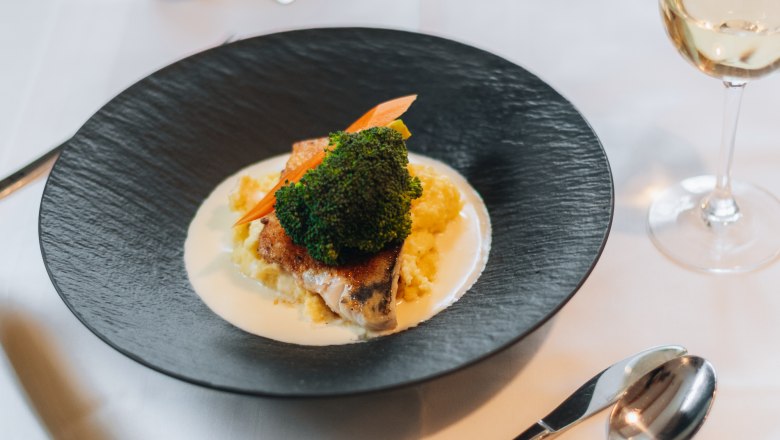 Ein elegant angerichtetes Gericht auf einem schwarzen Teller mit Gemüse und Sauce, daneben ein Glas Weißwein auf einem gedeckten Tisch.