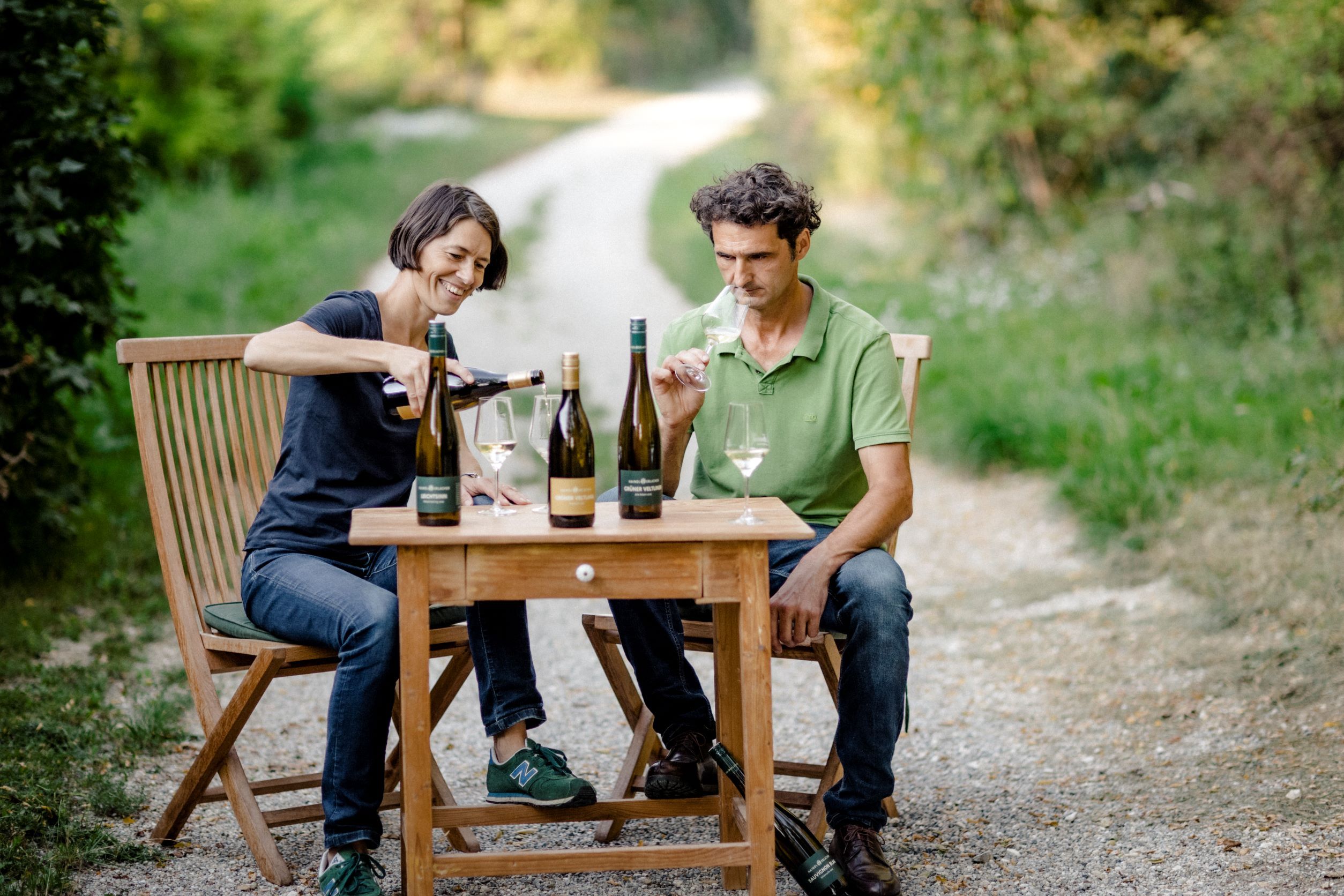 Zwei Personen sitzen an einem Holztisch im Freien und verkosten Wein.