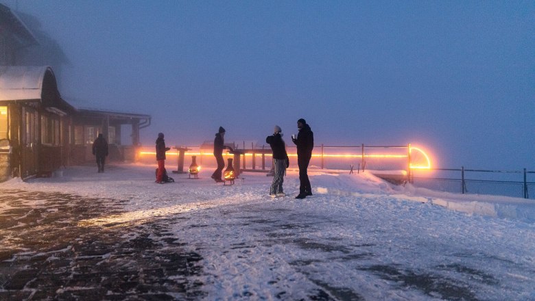 Bergadvent Rax, Advent, Raxalm-Bergstation, Winter, Rax-Seilbahn, Region Semmering-Rax, Wiener Alpen in Niederösterreich, © Wiener Alpen/Christian Kremsl Menschen stehen auf verschneiter Bergterrasse im Nebel, beleuchtet von warmen Lichtern bei Dämmerung.