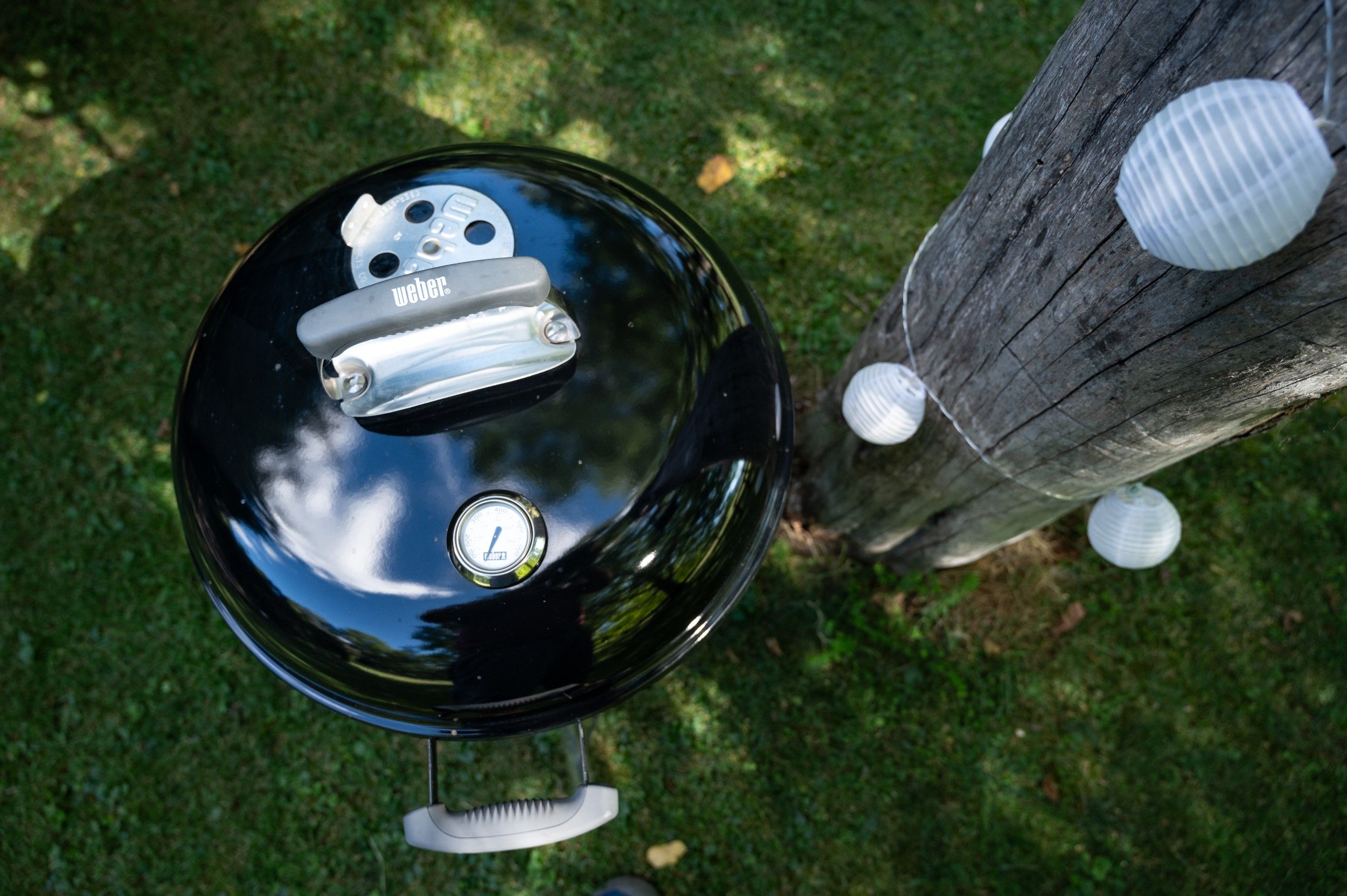 Ein Kugelgrill neben einem Baumstamm mit Lichterkette im Garten.