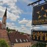 Blick auf ein Schild mit der Aufschrift 'Golden Star Premium Apartments' und 'Goldener Stern' vor einem Kirchturm und Dächern.
