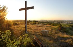 Ein Kreuz auf einem Hügel bei Sonnenuntergang mit Blick auf eine weite Landschaft und ein Dorf im Hintergrund.