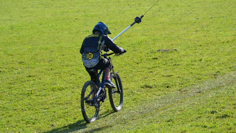 Ein Mountainbiker wird mit einem Schlepplift auf einer gr&uuml;nen Wiese gezogen.