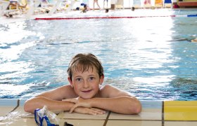 Ein Junge lehnt am Beckenrand eines Hallenbads, lächelt und hat eine Schwimmbrille neben sich.