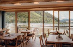 Innenansicht eines Gasthofes mit großen Fenstern und Blick auf eine Brücke und bewaldete Hügel.