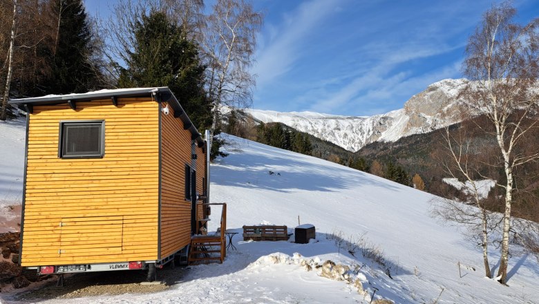 Kleines Holzhaus auf schneebedecktem H&uuml;gel mit Berglandschaft im Hintergrund.
