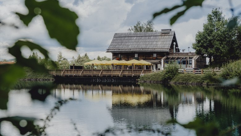 Ein rustikales Holzhaus mit gelben Sonnenschirmen spiegelt sich in einem ruhigen See, umgeben von grüner Natur.