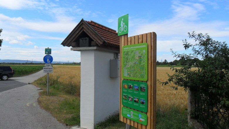 Radstartplatz-Tafel in Katzelsdorf mit Wegweiser und Landschaft im Hintergrund.
