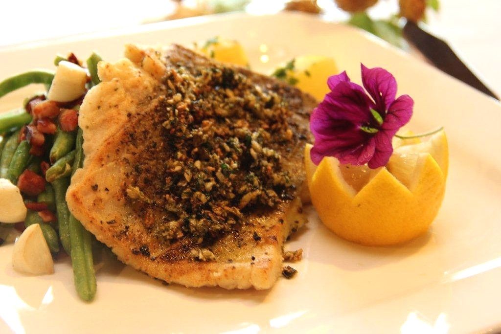 Ein Teller mit gebratenem Fischfilet, dekoriert mit einer Blume und einer Zitronenscheibe, serviert mit grünen Bohnen und Speck.
