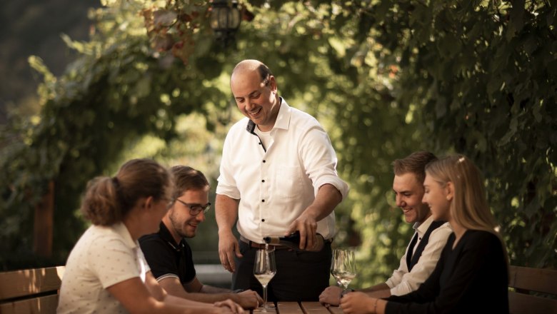 Ein Mann schenkt Wein für vier Gäste an einem Tisch im Freien ein, umgeben von grünen Weinreben.