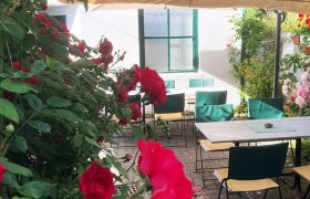 Ein gemütlicher Außenbereich eines Cafés mit Tischen, Stühlen und roten Rosen unter einem Sonnenschirm.
