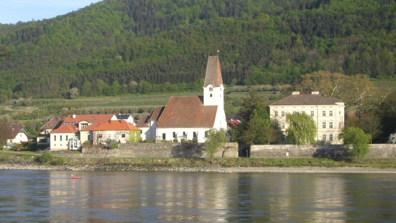 Pfarrkirche Hofarnsdorf mit umliegenden Gebäuden und bewaldetem Hügel im Hintergrund, aufgenommen von der Donau aus.