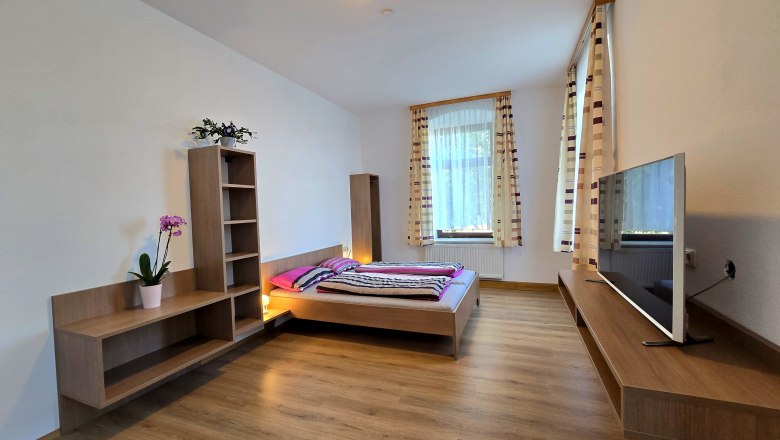 Apartment 2 - Schlafbereich, © Apartment Bauer Zur Alten Schule Modernes Schlafzimmer mit Holzboden, Doppelbett, Fernseher und Regalen. Große Fenster mit gestreiften Vorhängen.