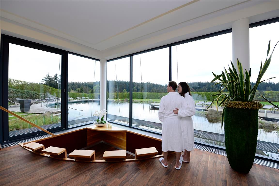 Ein Paar in Bademänteln steht in einem modernen Wellnessbereich mit Blick auf einen Teich.