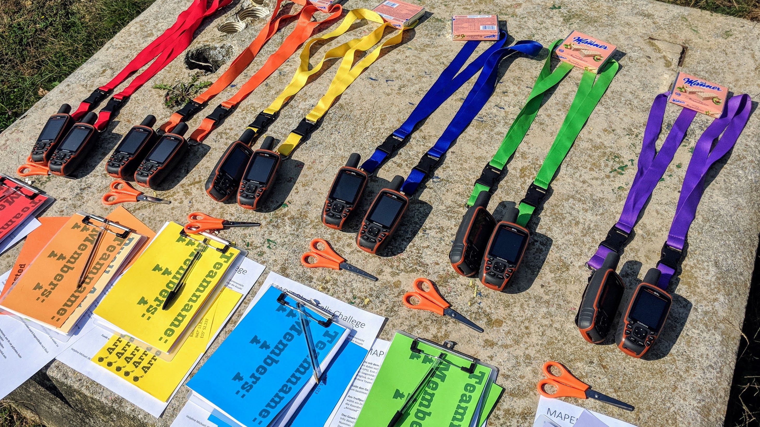 Bunt vorbereitete Teambuilding-Station mit GPS-Geräten, farbigen Team-Bändern und Aufgabenmappen für eine spannende Outdoor-Challenge in der Natur.