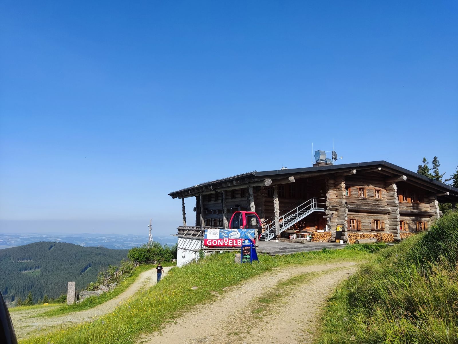 Eine Berghütte aus Holz mit einem roten Fahrzeug davor, umgeben von grünen Wiesen und einem klaren blauen Himmel.