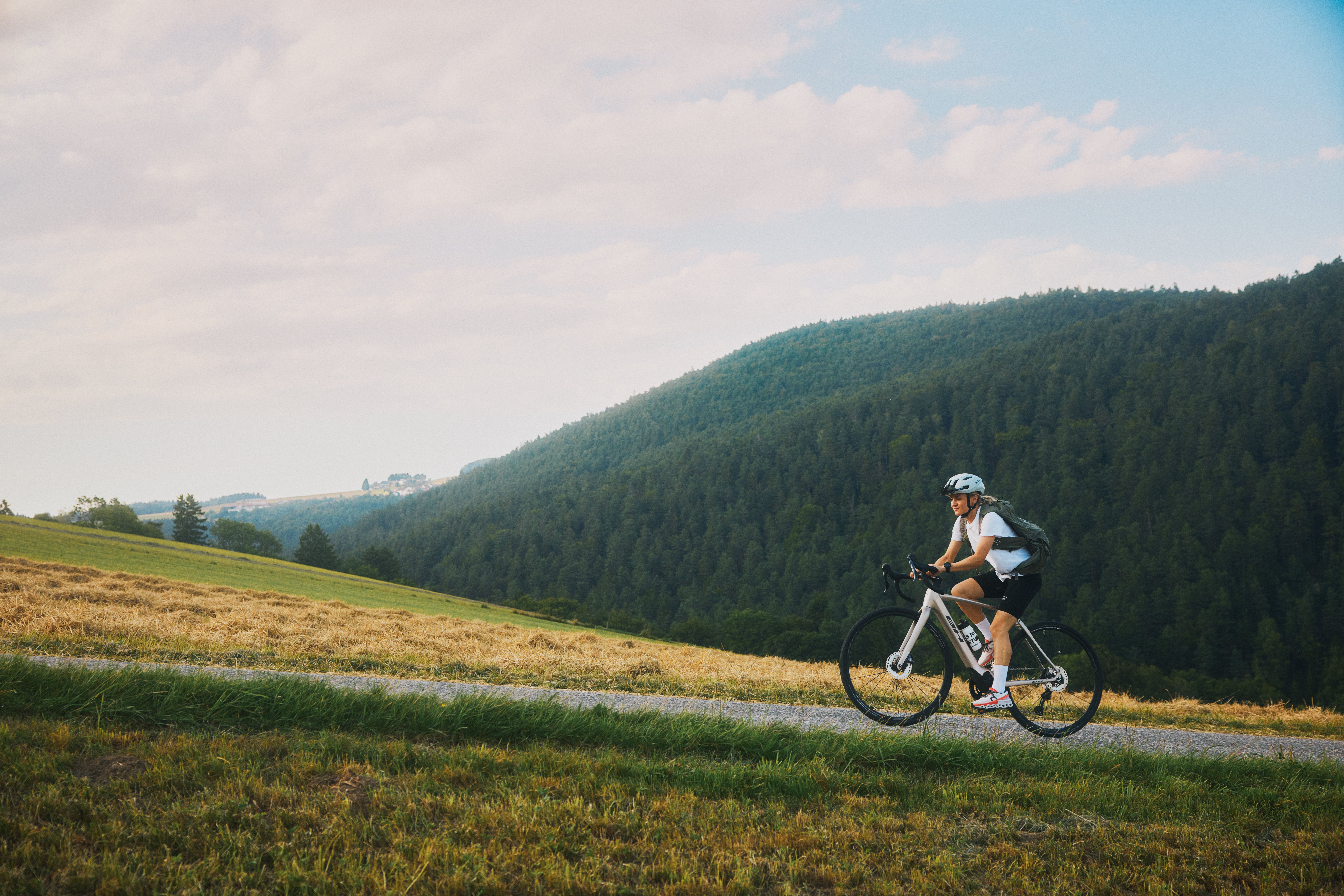 Eine Radfahrerin fährt auf einem schmalen, steigenden Weg, im Hintergrund bewaldete Hügel.