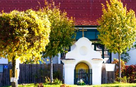 Ein kleines, wei&szlig;es Kapellengeb&auml;ude mit einem Kreuz auf dem Dach, umgeben von herbstlich gef&auml;rbten B&auml;umen.
