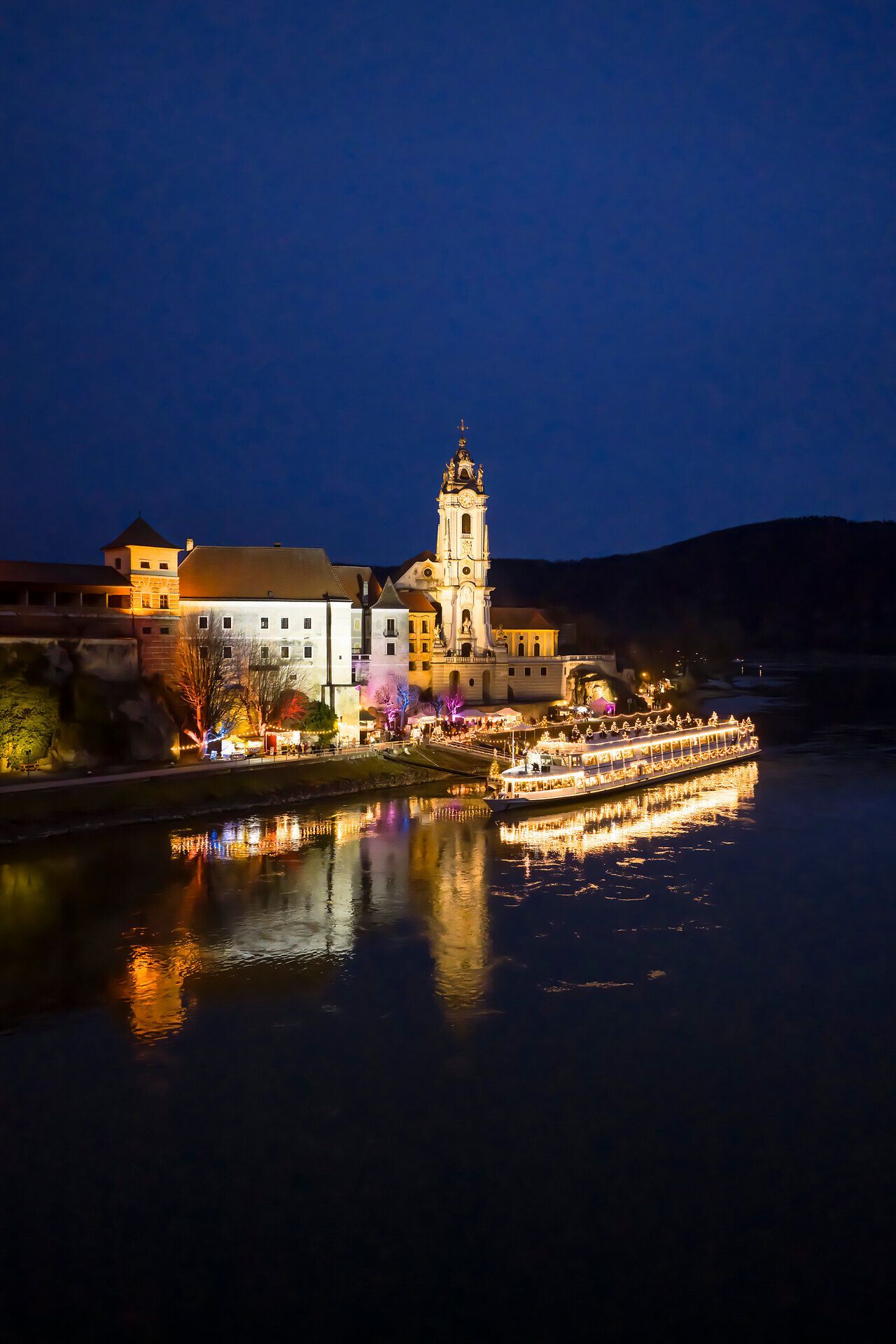 Nächtlicher Blick auf das festlich beleuchtete Dürnstein an der Donau, mit reflektierenden Lichtern, ruhiger Flusslandschaft und historischer Kirche im Advent.