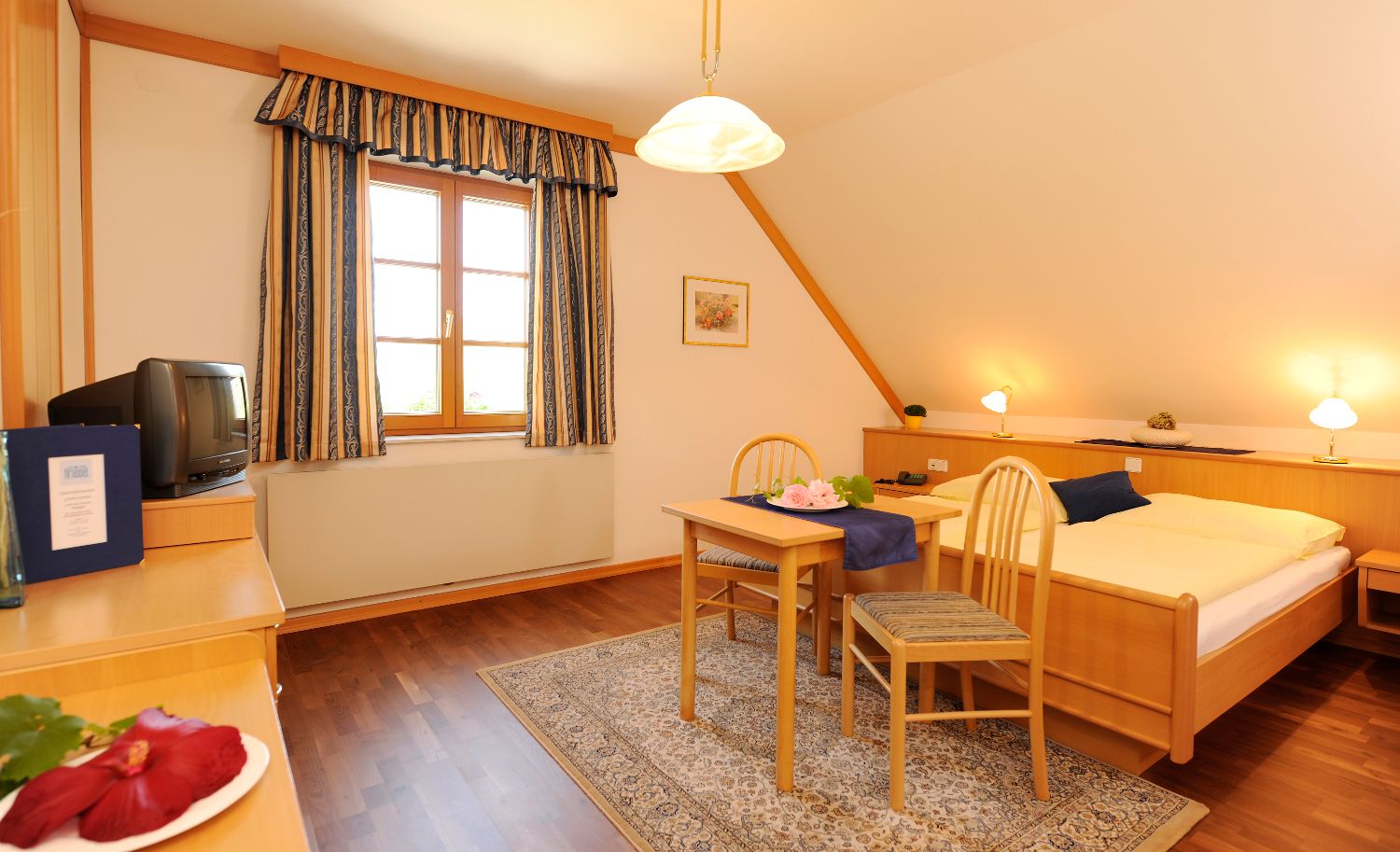 Gemütliches Gästezimmer mit Holzmöbeln, Doppelbett, kleinem Tisch und Stühlen, TV und Fenster mit Vorhängen.