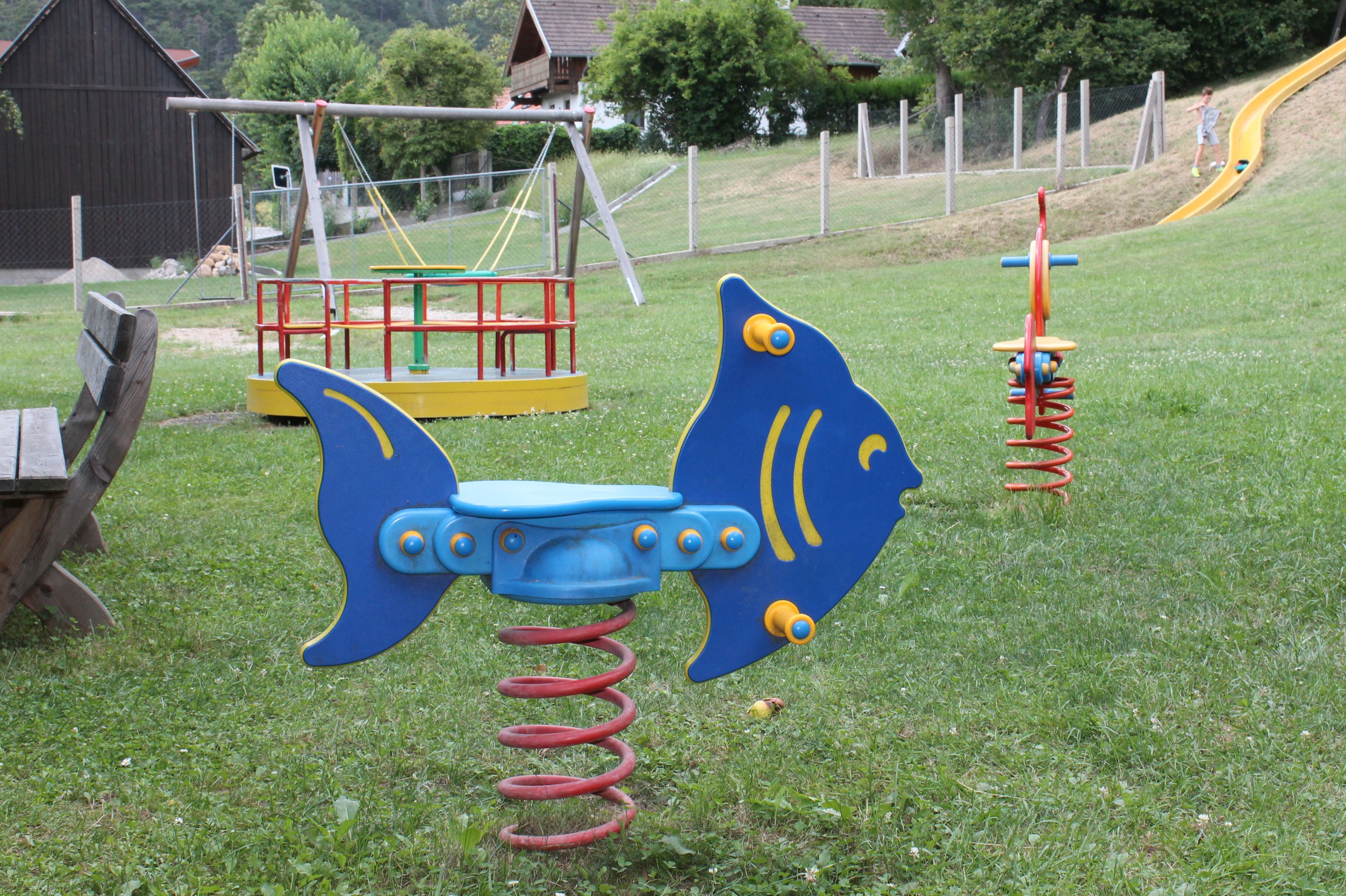 Spielplatz mit Wipptier in Fischform, Karussell und Rutsche.